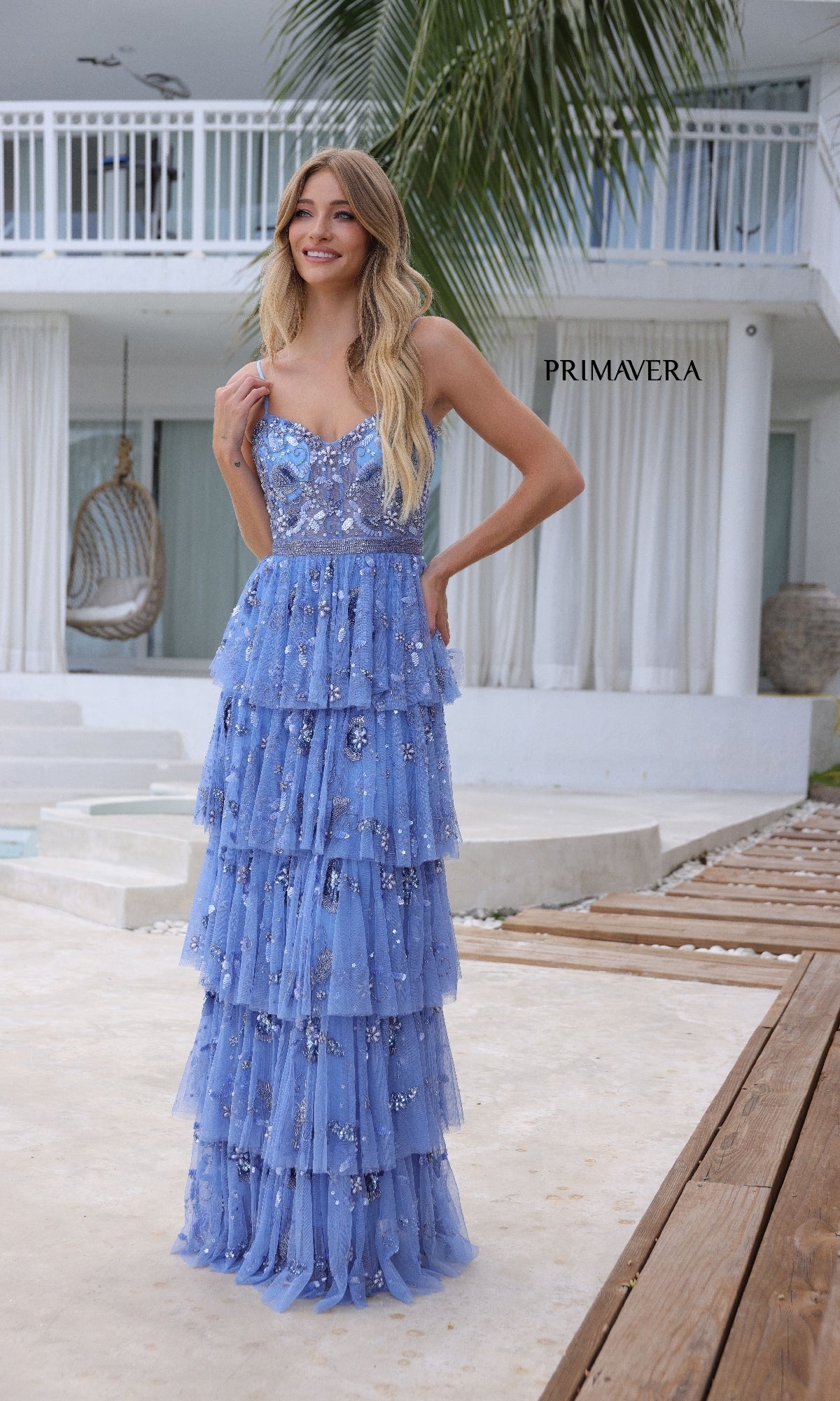 A woman stands outdoors in front of a modern white house, smiling at the camera in the Long Tiered Sequin Prom Dress: Primavera 4341.